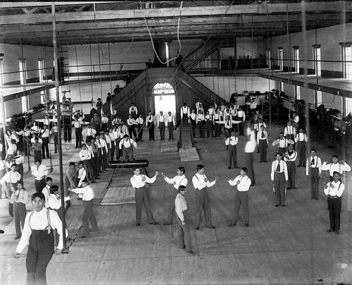 Carlisle_Students_in_School_Uniform_Exercising_Inside_Gymnasium;_Some_with_Indian_Clubs,_Others_with_Gymnastic_Equipment;_Non-Native_Group_Watching_1879